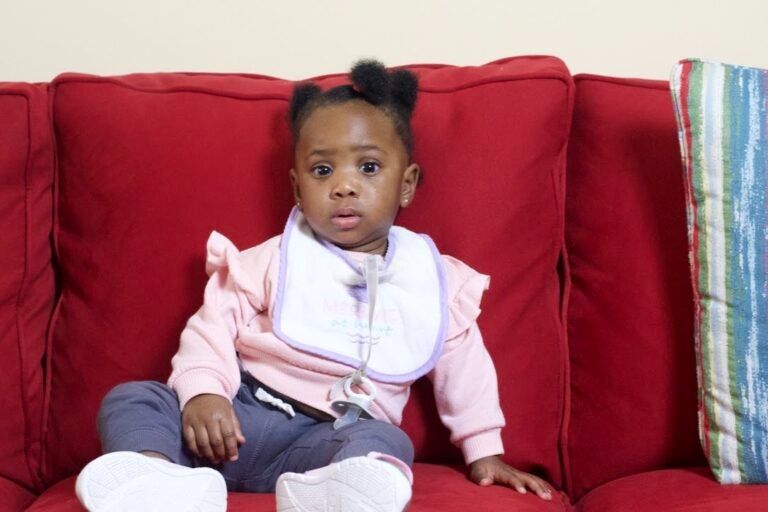A baby wearing a pink shirt and white bib sits on a bright red couch.