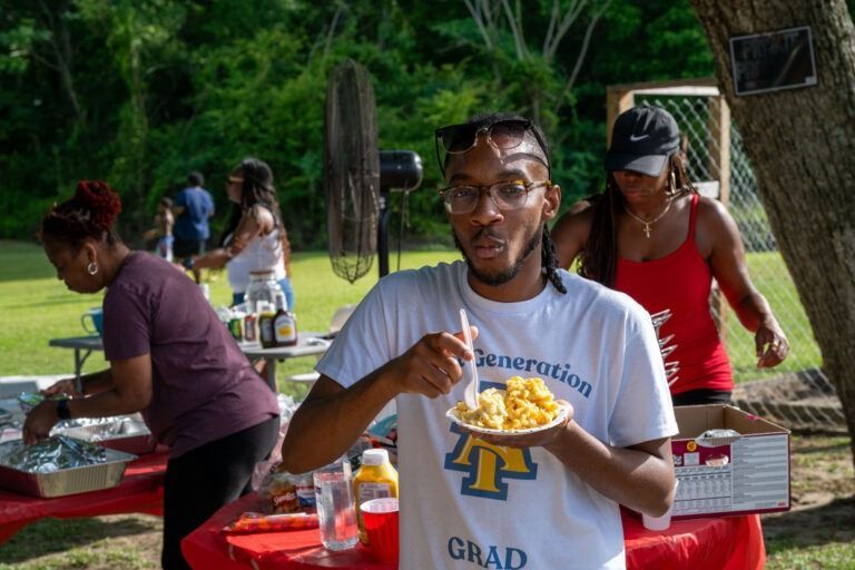 A person in a white t-shirt eats from a paper plate at an outdoor park gathering with others preparing food in the back.