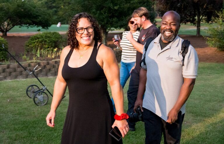 Four people walk outside on a grass lawn during the day; a woman in a black dress leads with a smile.