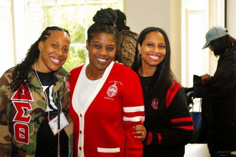 Three smiling women pose together indoors, wearing red and black apparel with Greek letters, with one person in the back.