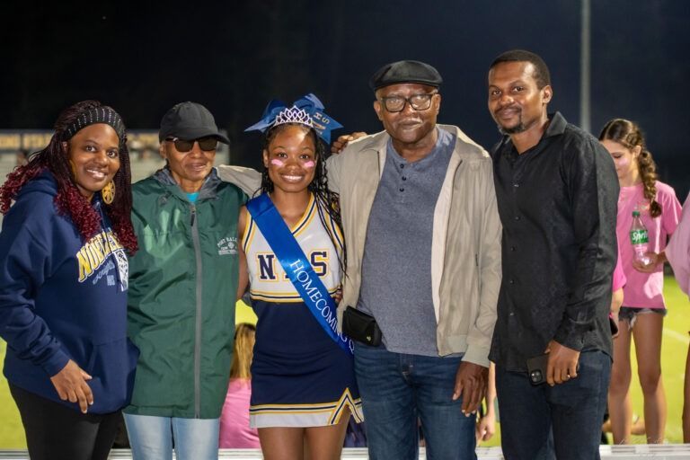A smiling cheerleader wearing a sash stands with four adults outdoors at night on a grassy field.