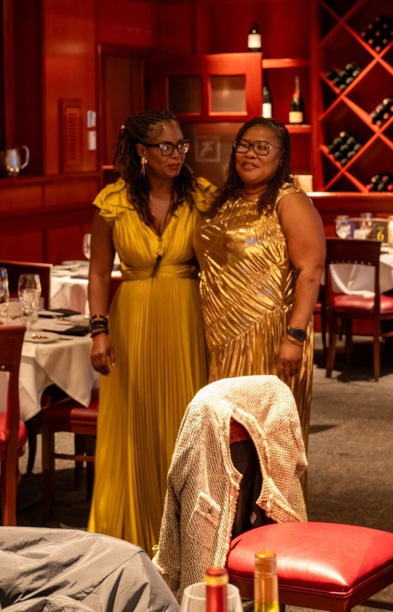 Two women in gold and yellow dresses standing in a red-toned restaurant with wine displays and set tables.