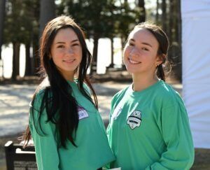 Two individuals smiling, wearing matching light green long-sleeved shirts with logos, standing outside near trees.