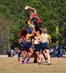 Rugby players in yellow and red jerseys perform a lineout lift, with one player jumping to catch the ball mid-air.