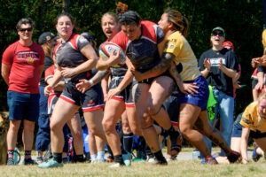 A rugby player in a dark jersey with a ball is tackled by an opponent in a yellow jersey, watched by people on a field.