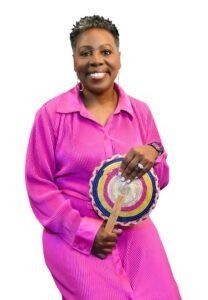 A smiling person wearing a bright pink long-sleeved shirt, holding a colorful round hand fan against a white background.