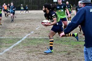 A rugby player in a green and black uniform runs with the ball on a field, while a coach watches from the sideline.