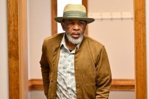 A person with a beard, wearing a hat and a brown jacket, stands in a hallway and looks slightly to the side.
