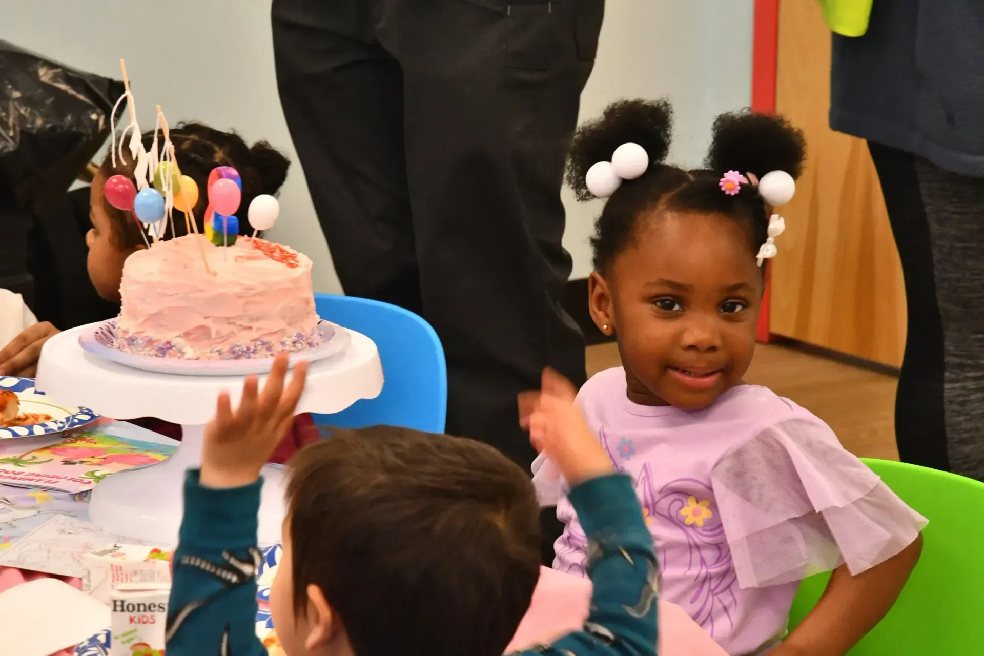 A young child with pigtails looks toward the camera beside a birthday cake with colorful balloons at a table.