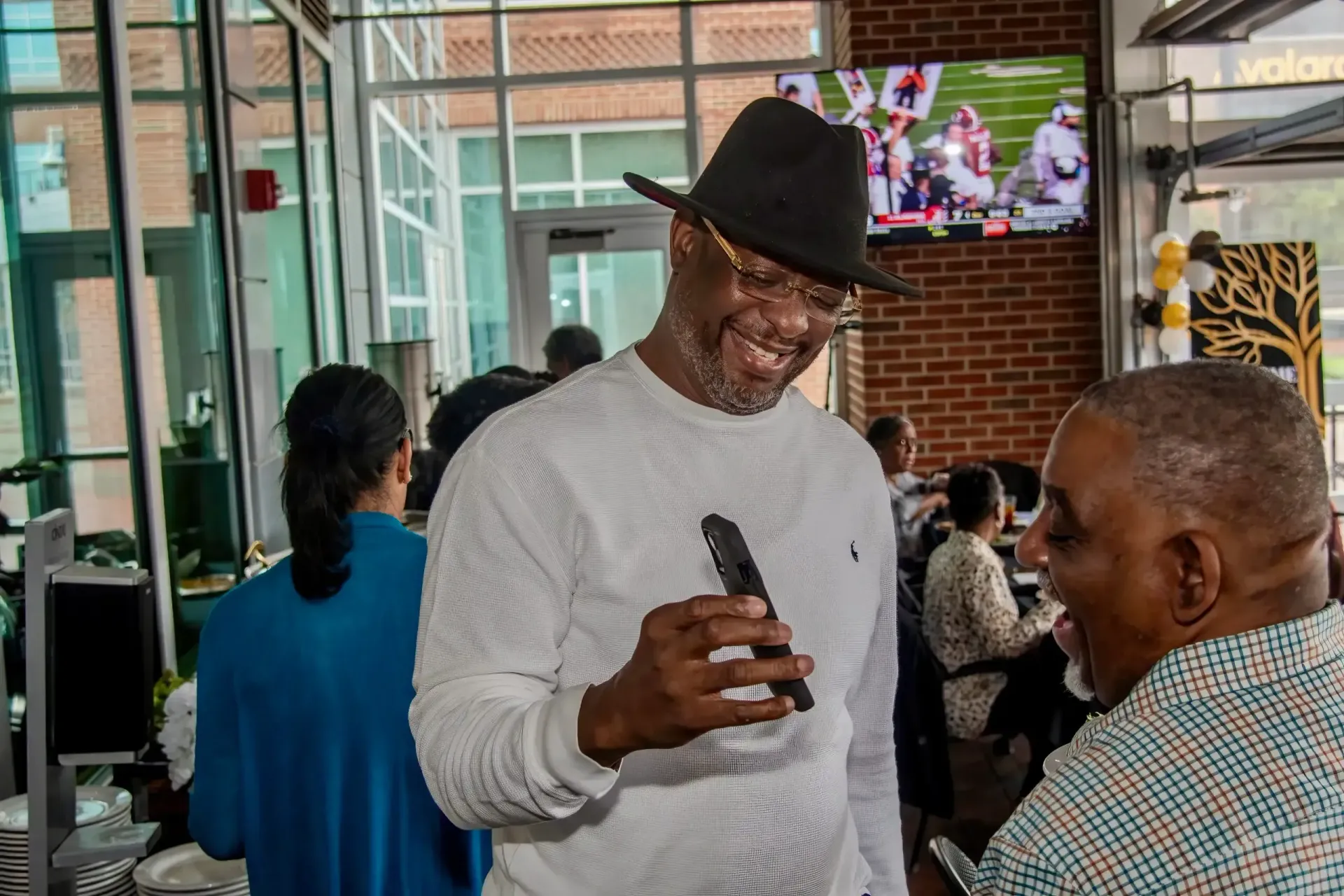 A man in a white sweater and black fedora holds a phone while smiling at another man during a social gathering indoors.