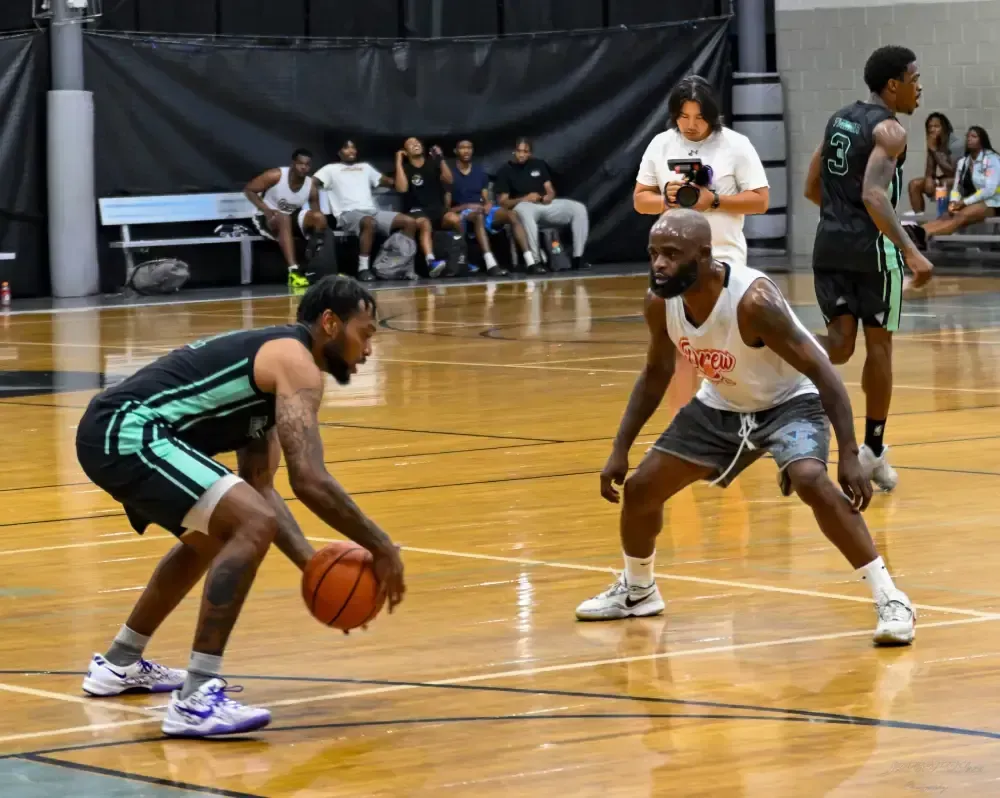 Two basketball players face each other on an indoor court, with one dribbling a ball while the other defends.