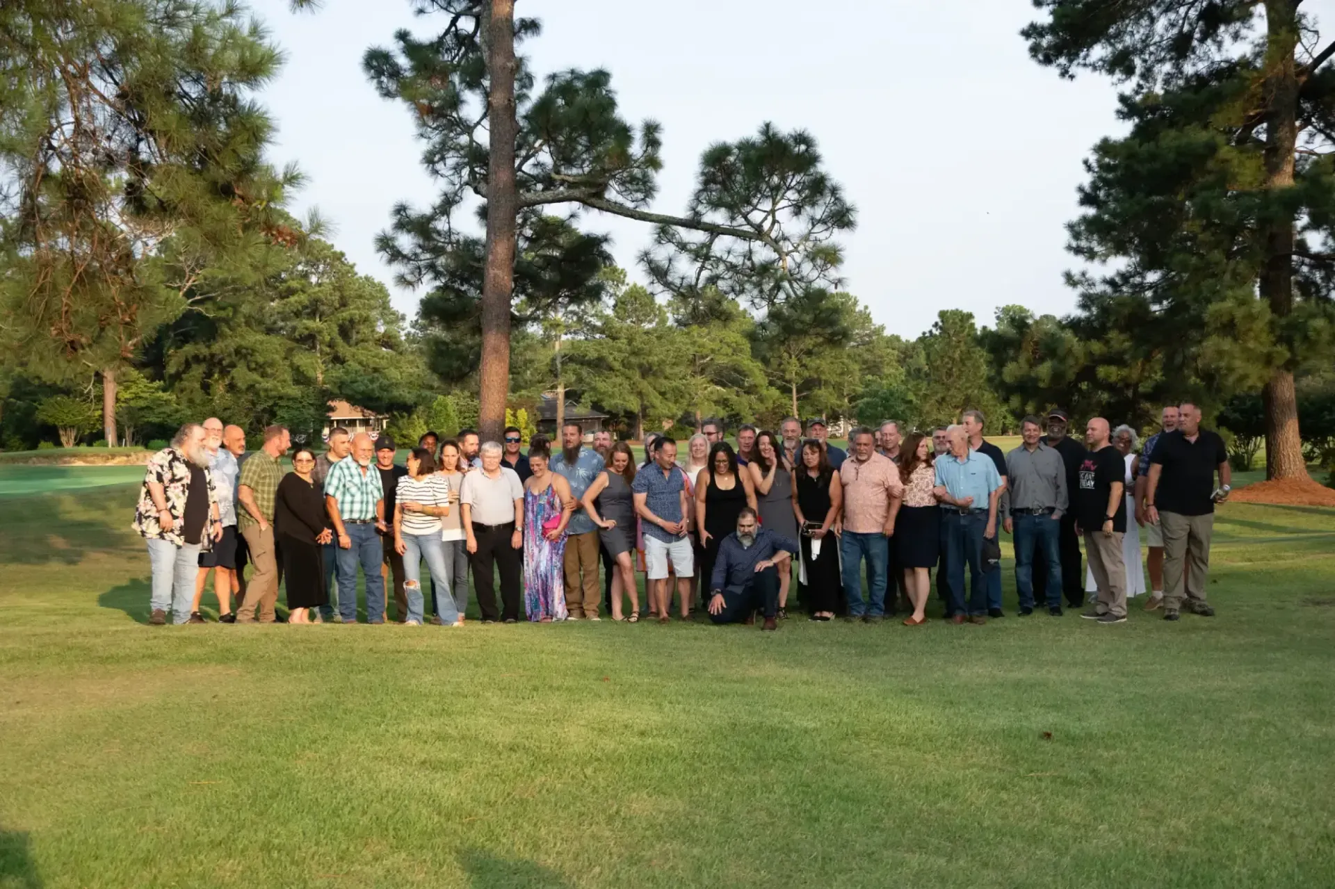 A large group of people poses for a photo on a grassy lawn surrounded by trees during an outdoor gathering.