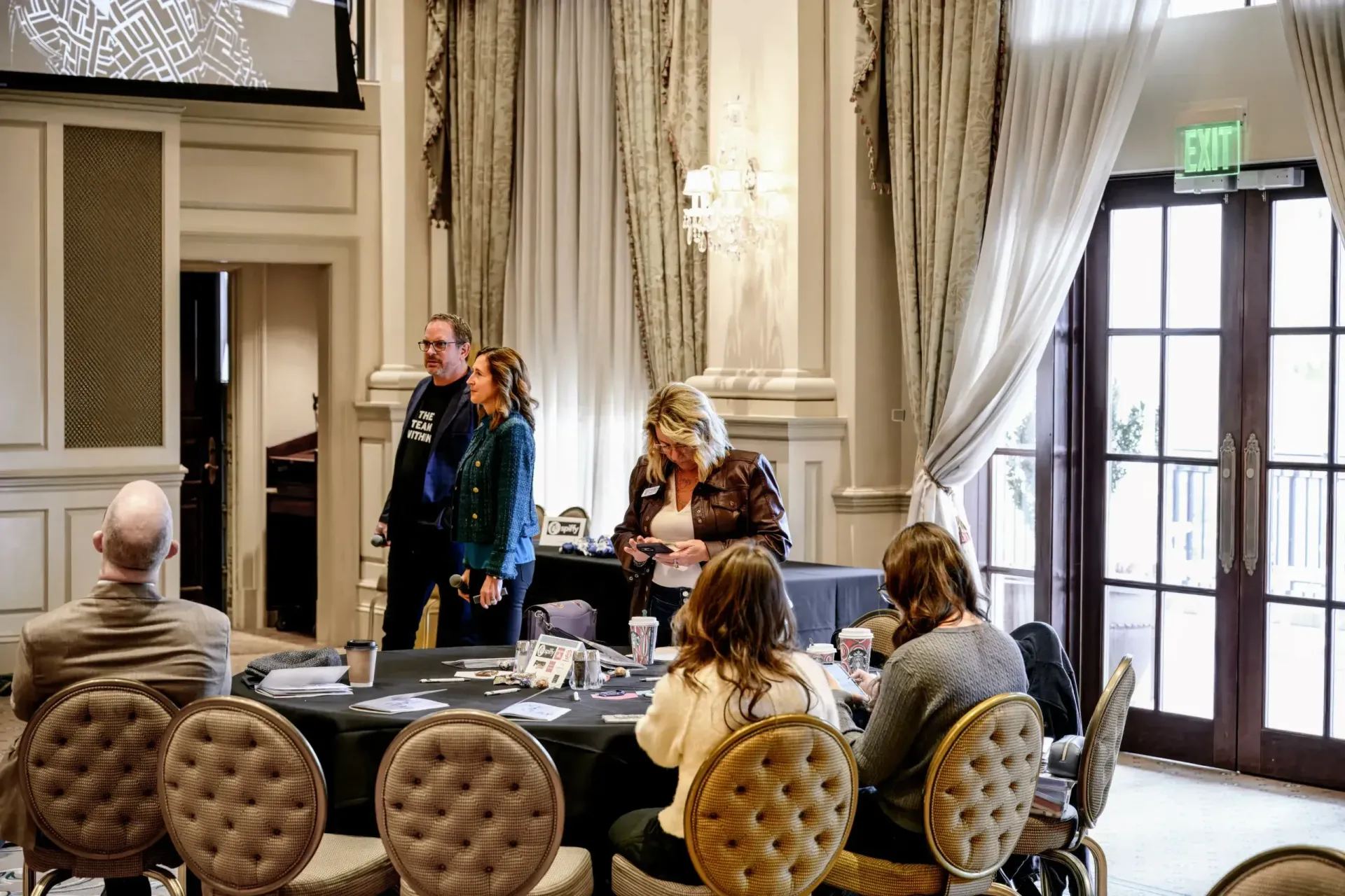 A group of people sitting at a round conference table in an ornate room with tall windows, curtains, and an exit sign.