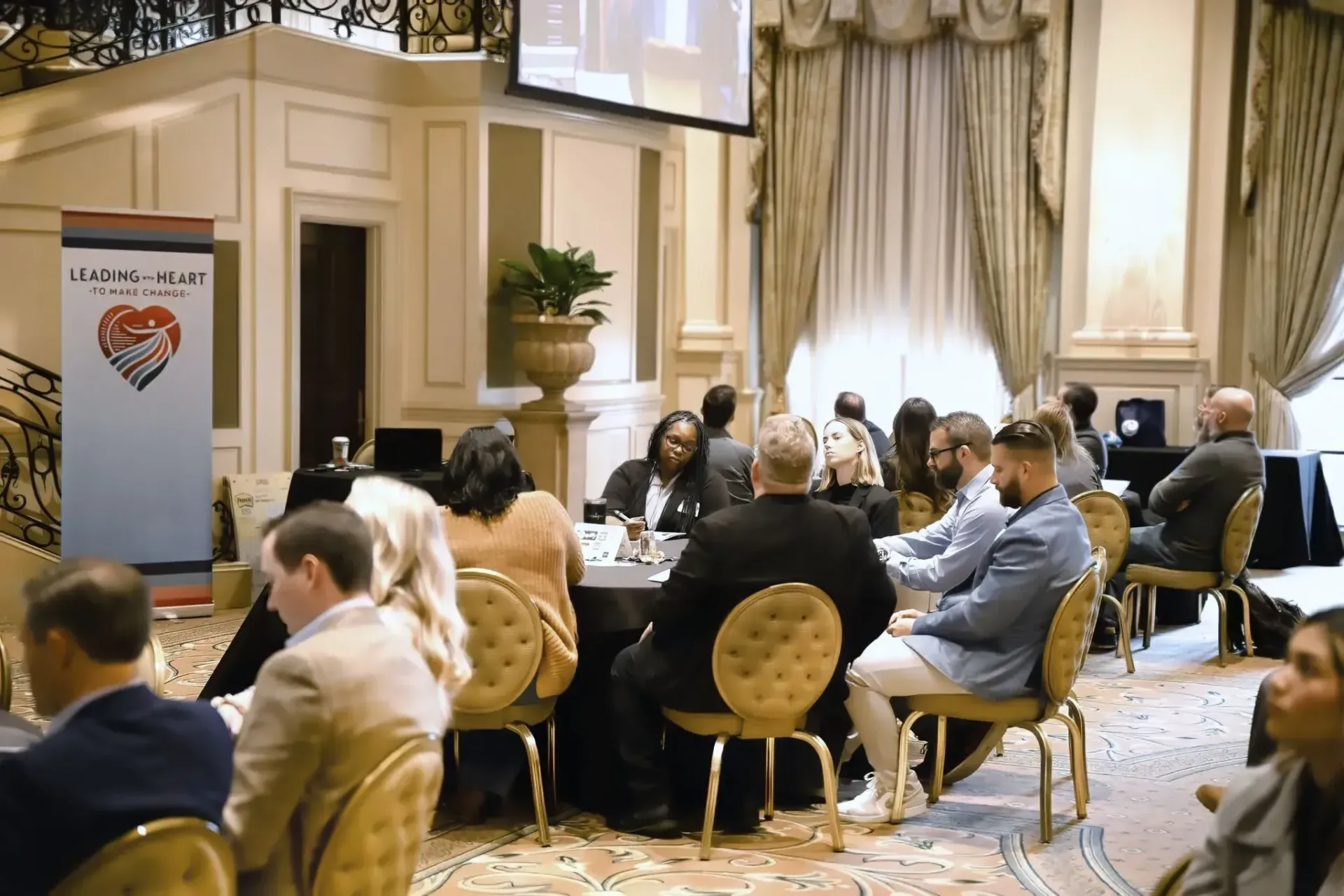 Attendees sit at tables in a ballroom with a banner displaying a heart-shaped logo featuring two hands joined in a clasp.