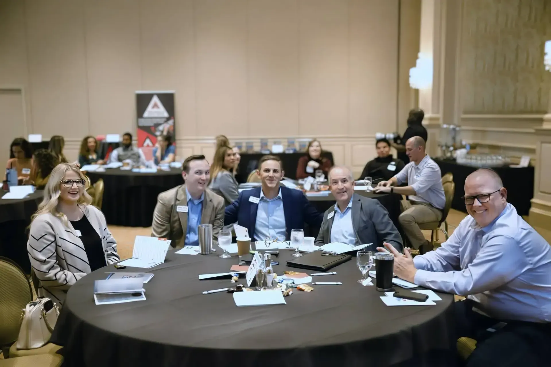 A group of people sit around a circular table in a conference room, smiling toward the camera during a professional event.