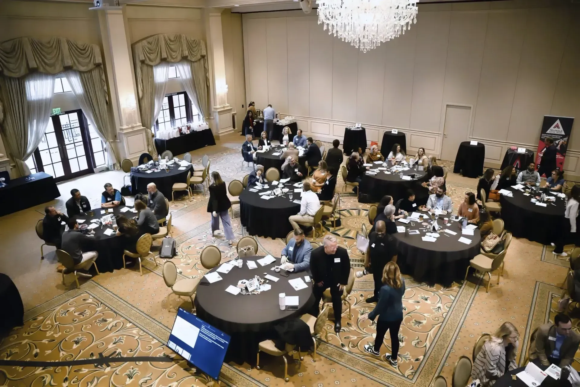 High-angle view of a well-lit banquet hall with people seated at round black tables for an event.