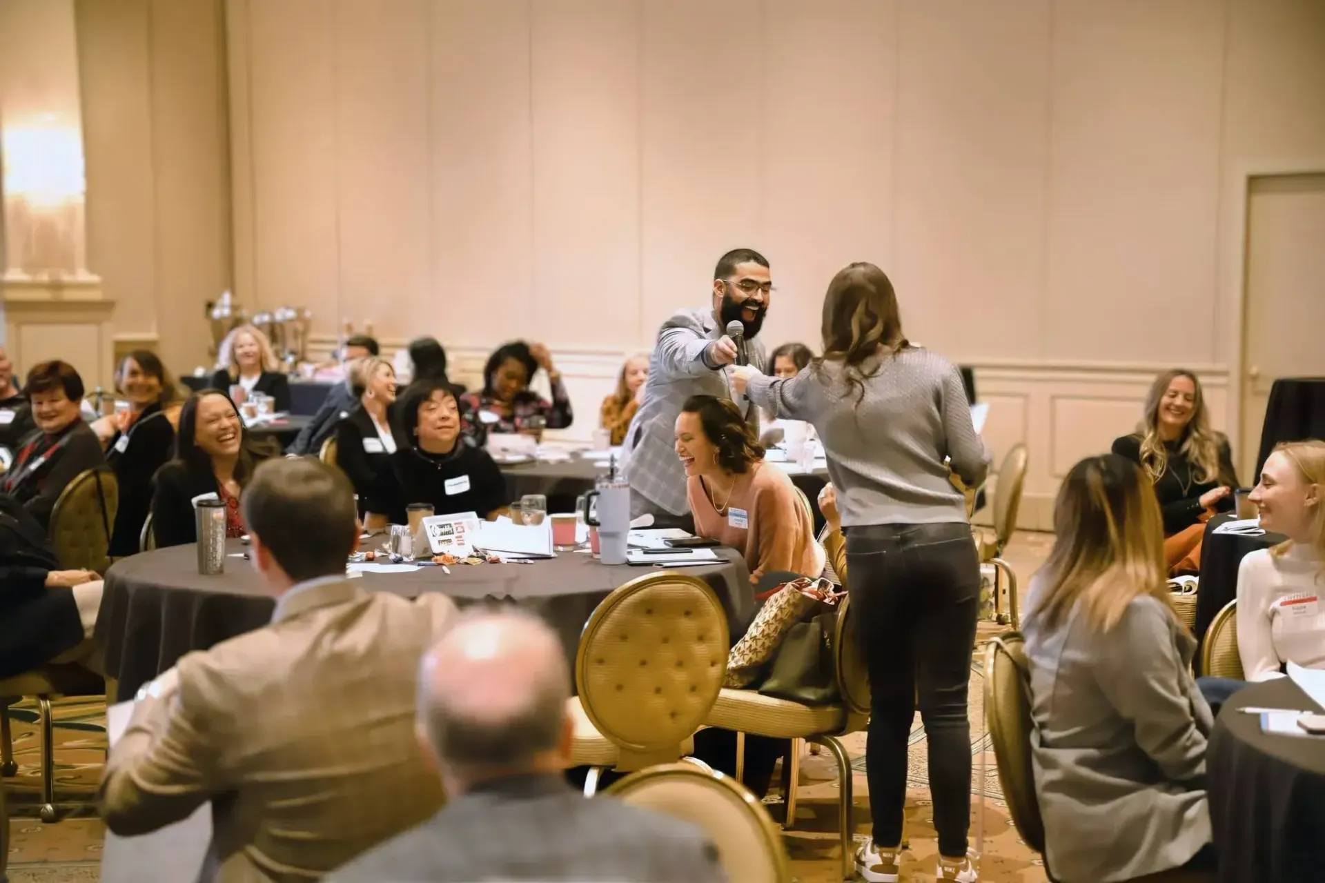 Attendees at a formal event sit at round tables while a speaker addresses the room with a microphone.