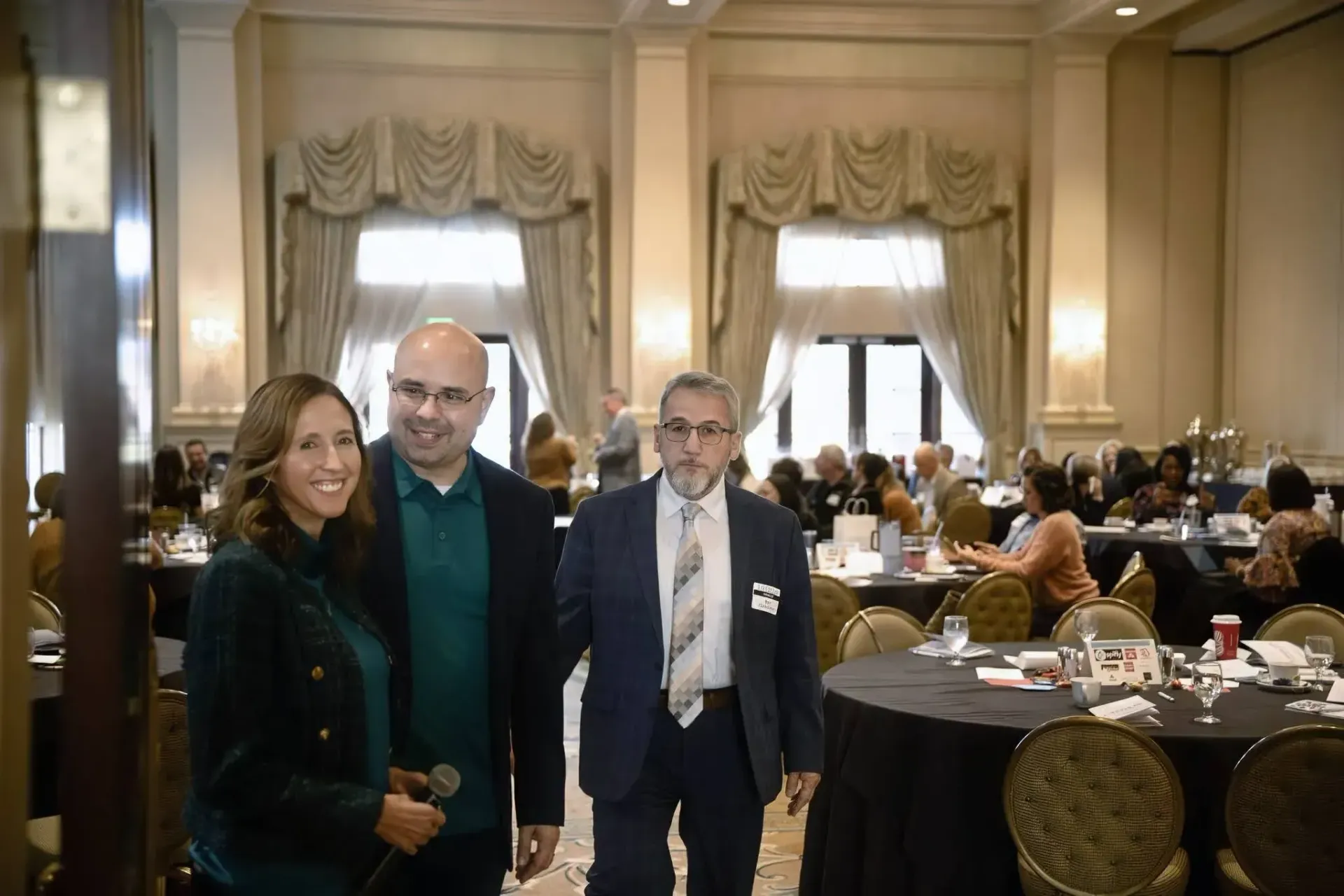 Three people stand in a banquet hall, two smiling toward the camera, in a room with draped windows and round tables.