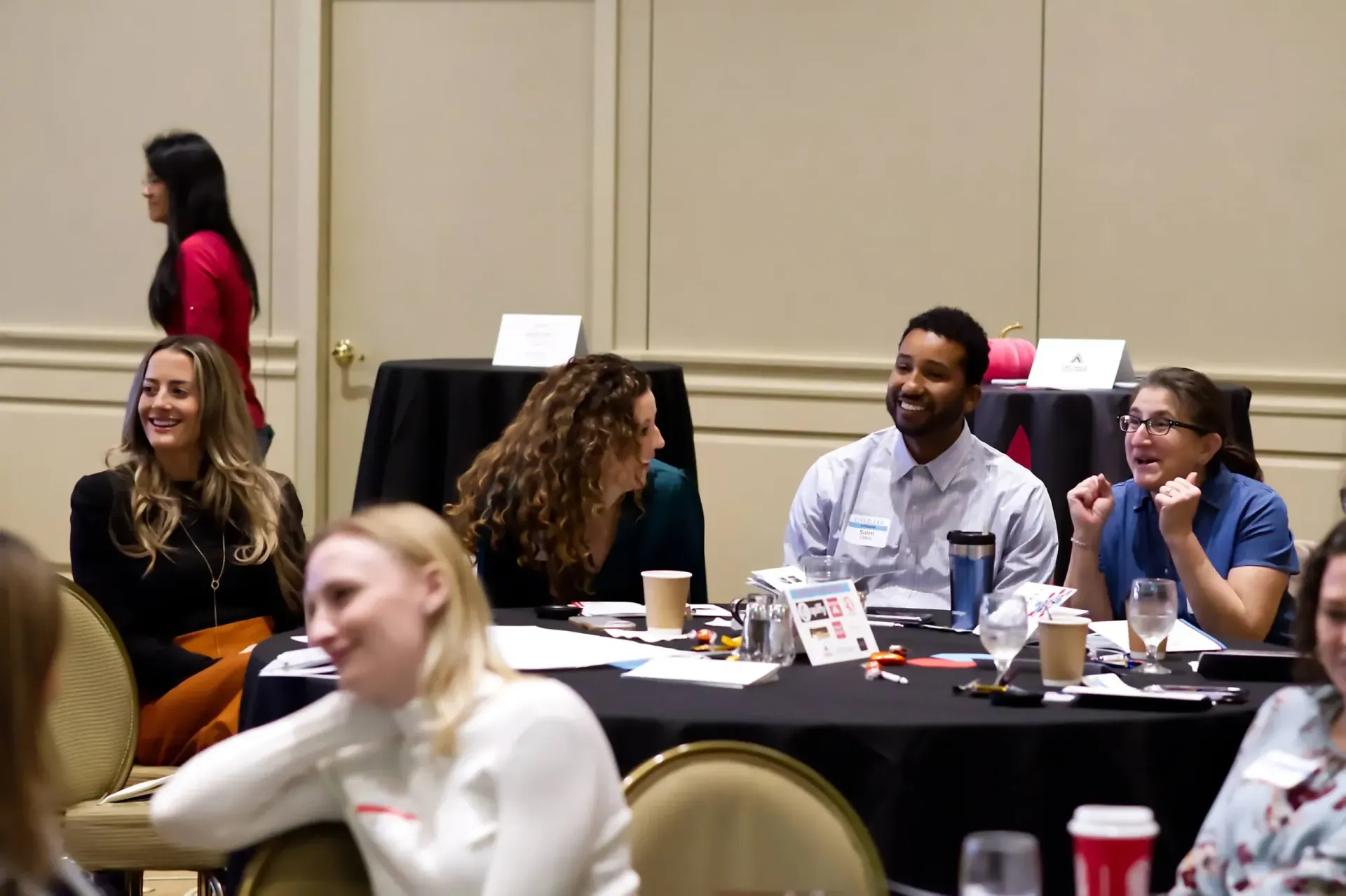 A group of people sit around a circular table in a conference room, listening and smiling during a presentation.