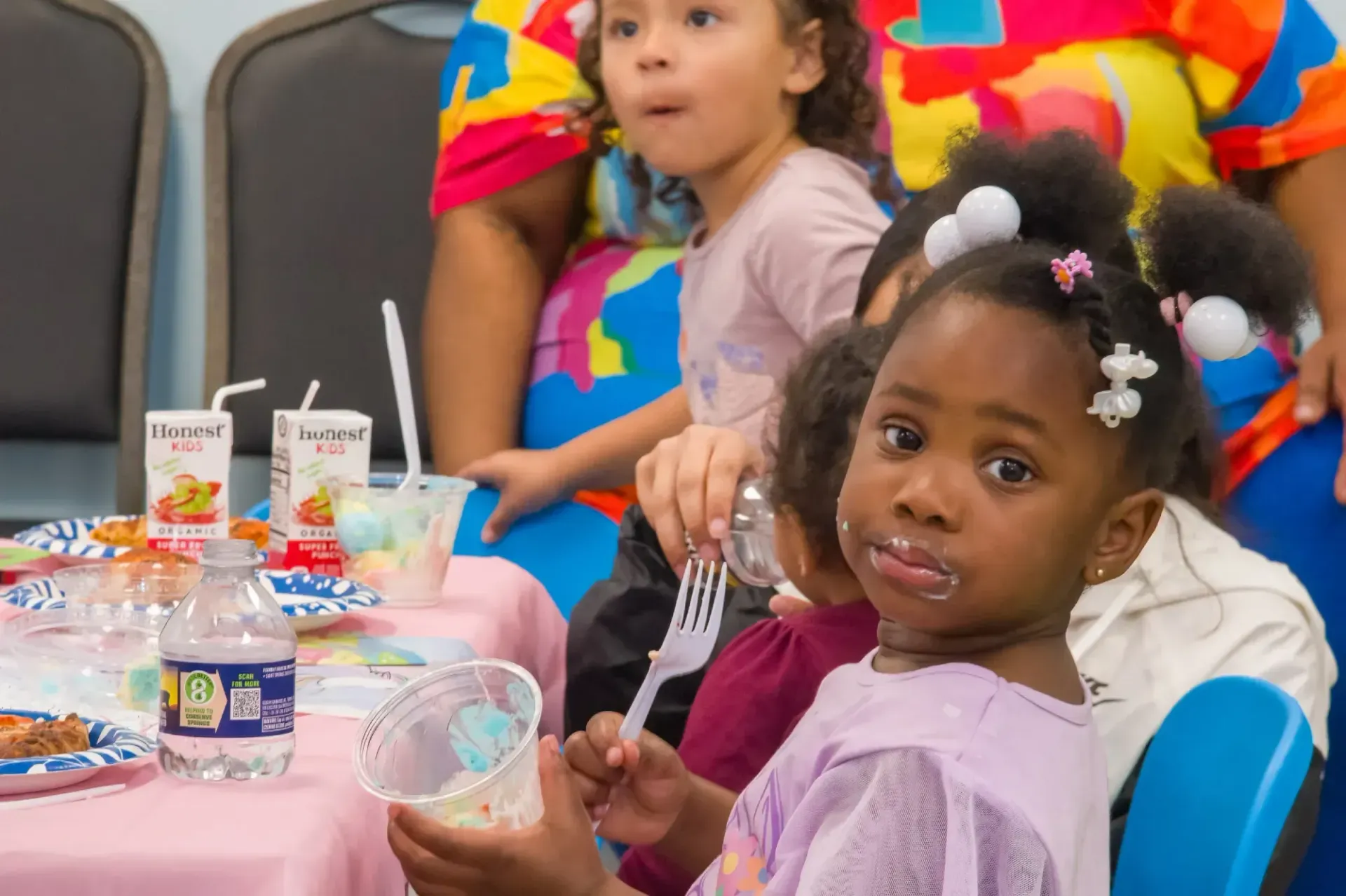 Children eating at a table with juice boxes and a water bottle, one looking at the camera with a smudge of food on mouth.