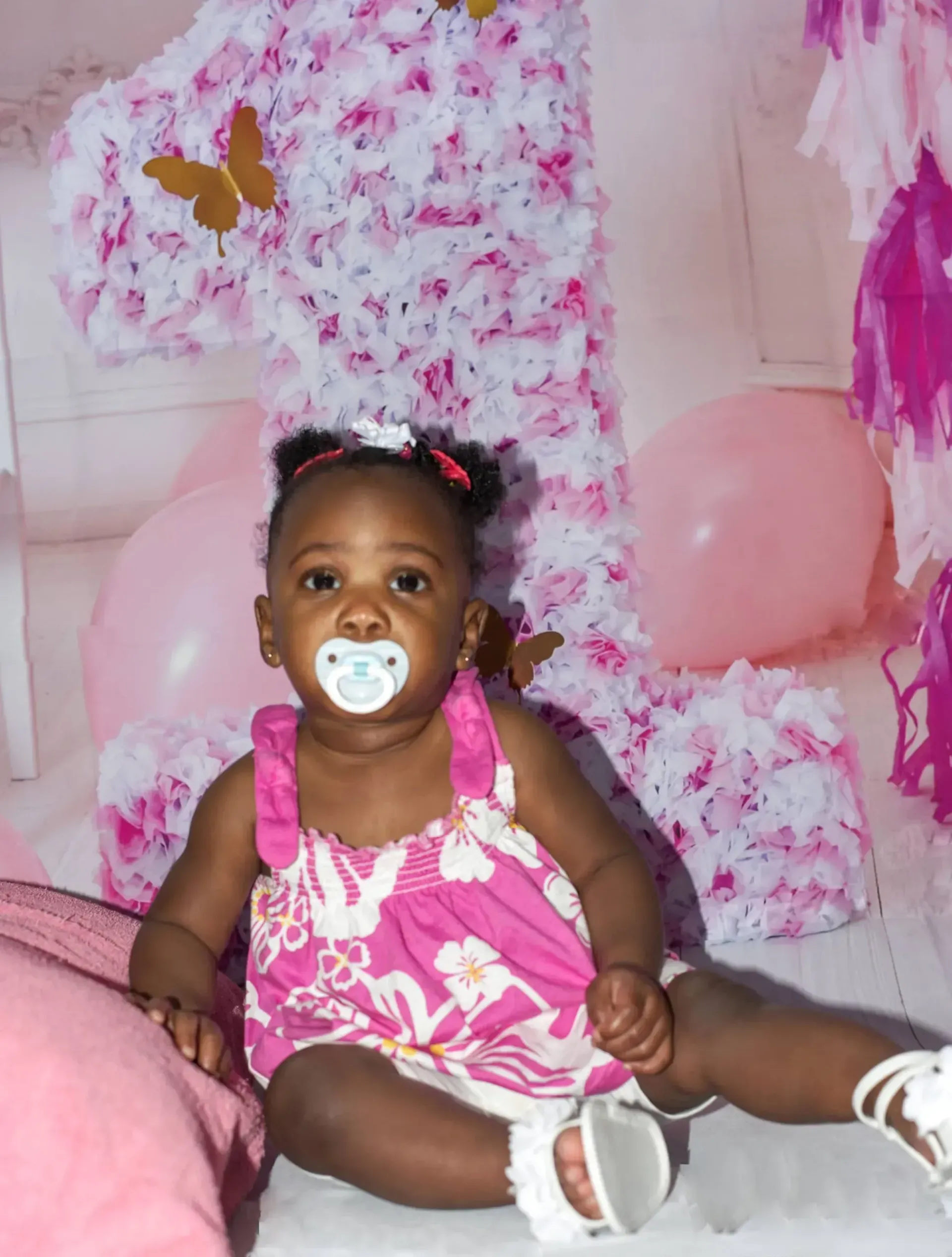 A baby wearing a floral pink top and white shoes sits in front of a large, floral