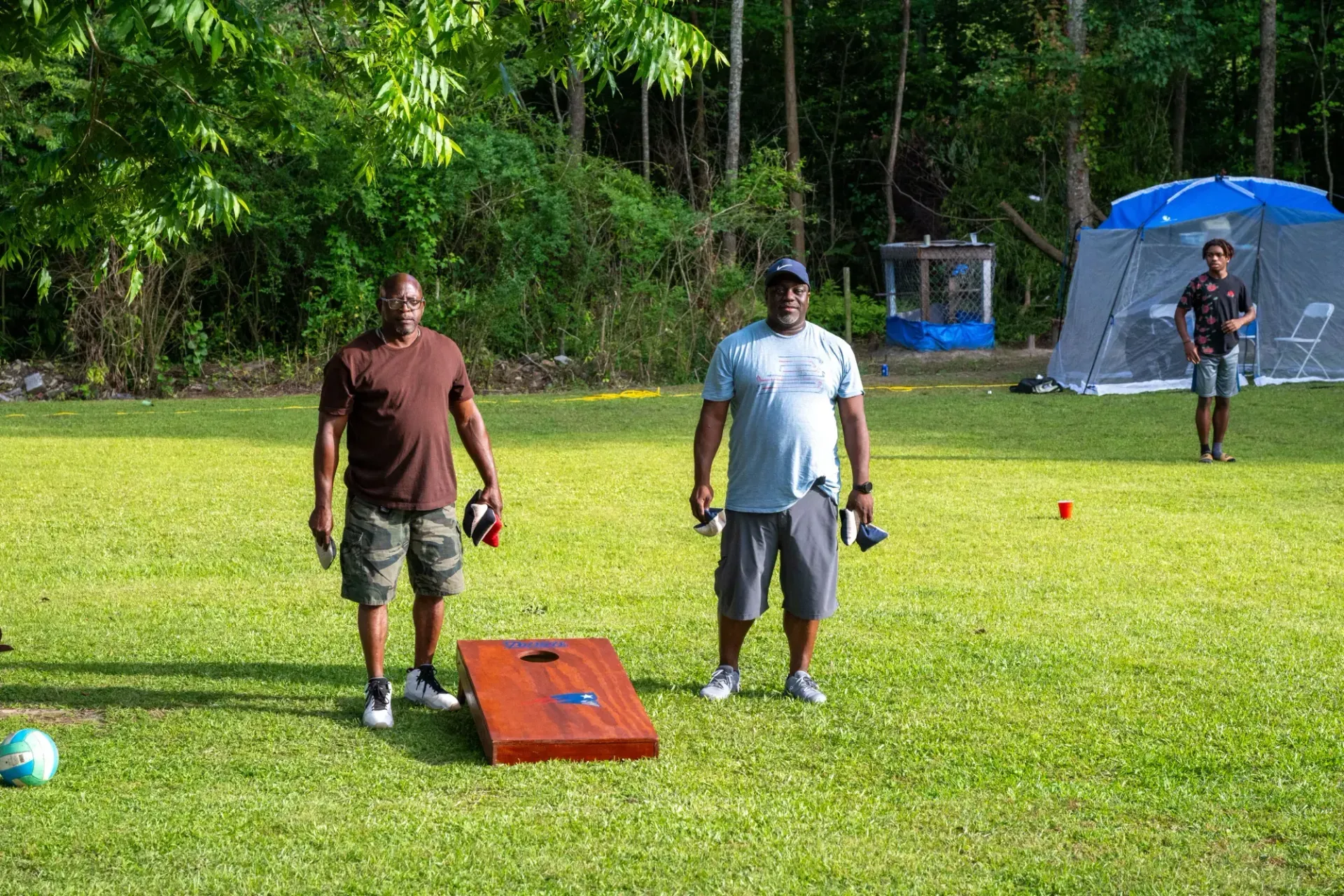 Two people stand on a grassy field by a cornhole board, while another person stands near a camping tent in the background.