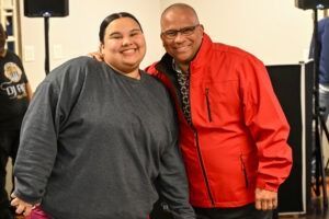 Two people standing side-by-side, smiling, indoors with audio equipment in the background.
