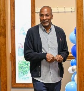 A person with glasses, a button-down shirt, and a navy cardigan standing in a room decorated with blue and white balloons.