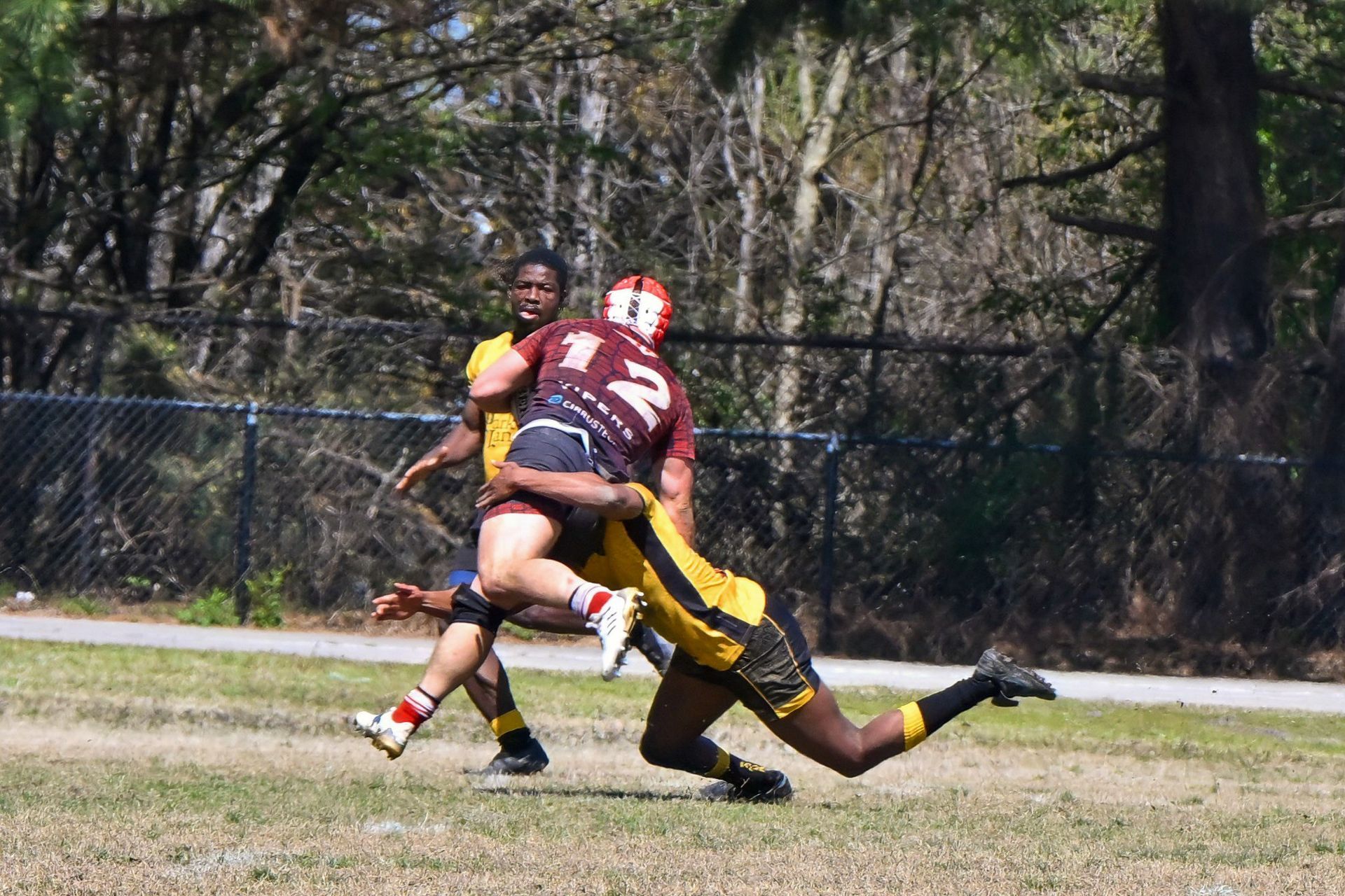 A rugby player in a maroon jersey is being tackled by an opponent in a yellow and black uniform on a grassy field.