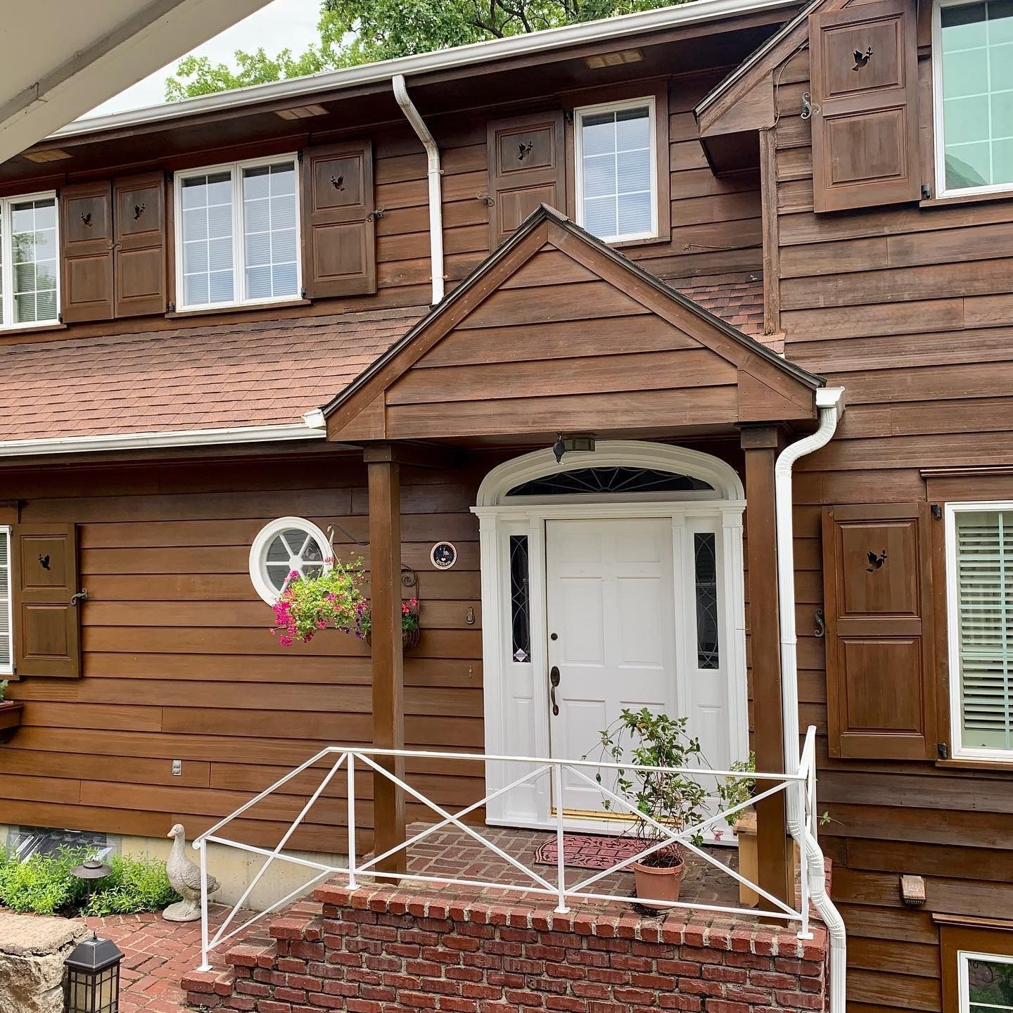 A brown, shingled two-story house with white-framed windows, dark shutters, and a white front door under a small porch.