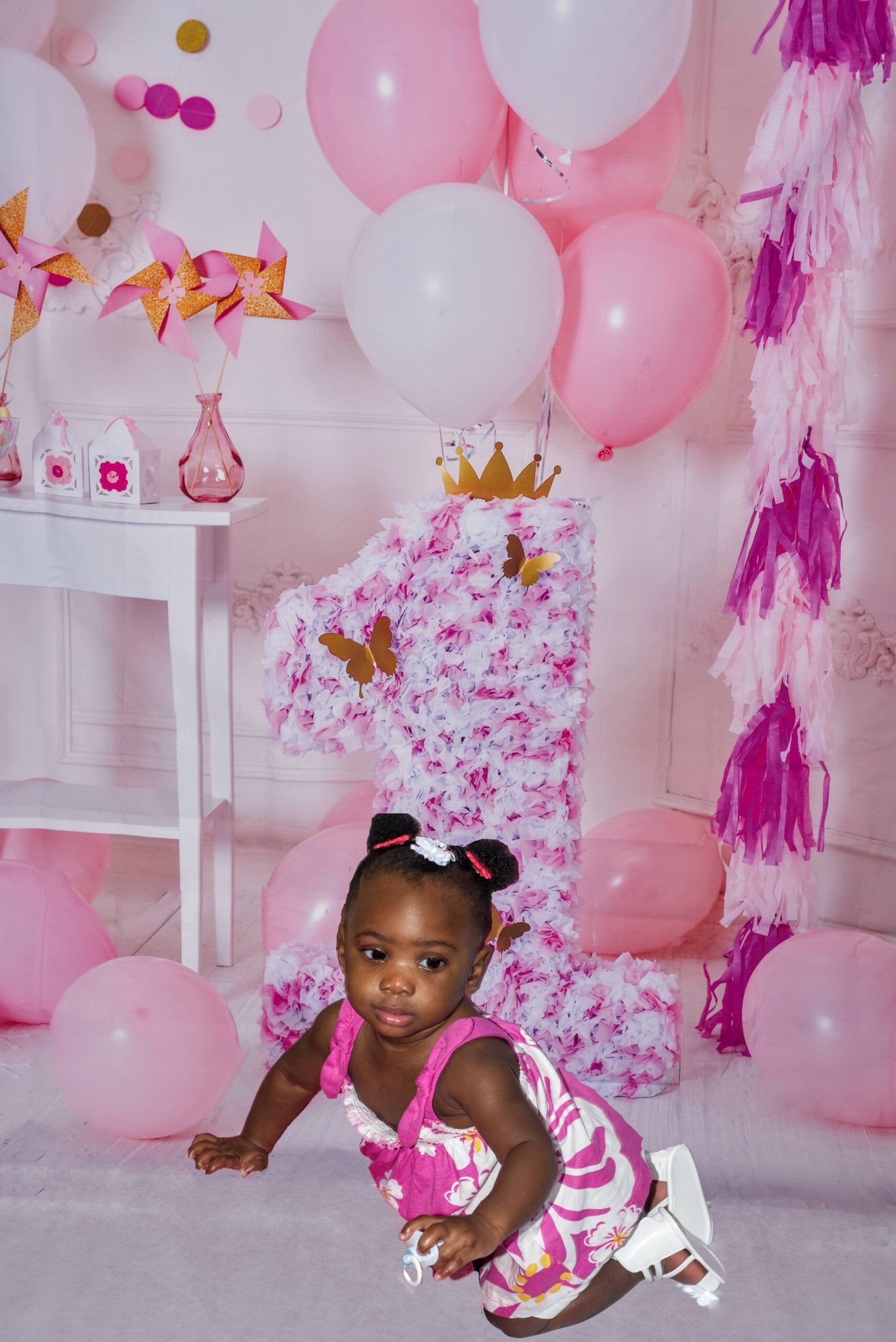 A toddler in a pink floral dress sits on the floor in front of a large floral-covered number one for a birthday celebration.
