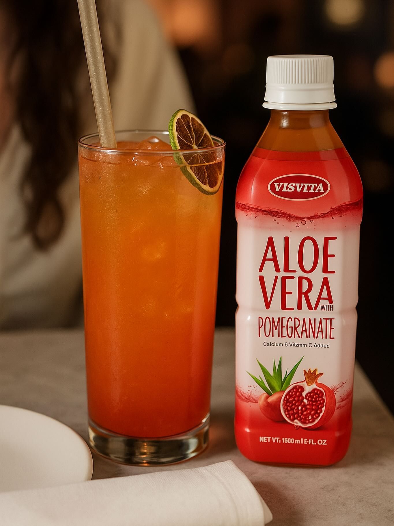 Glass of orange drink with straw and lime slice, next to a bottle of aloe vera pomegranate juice.