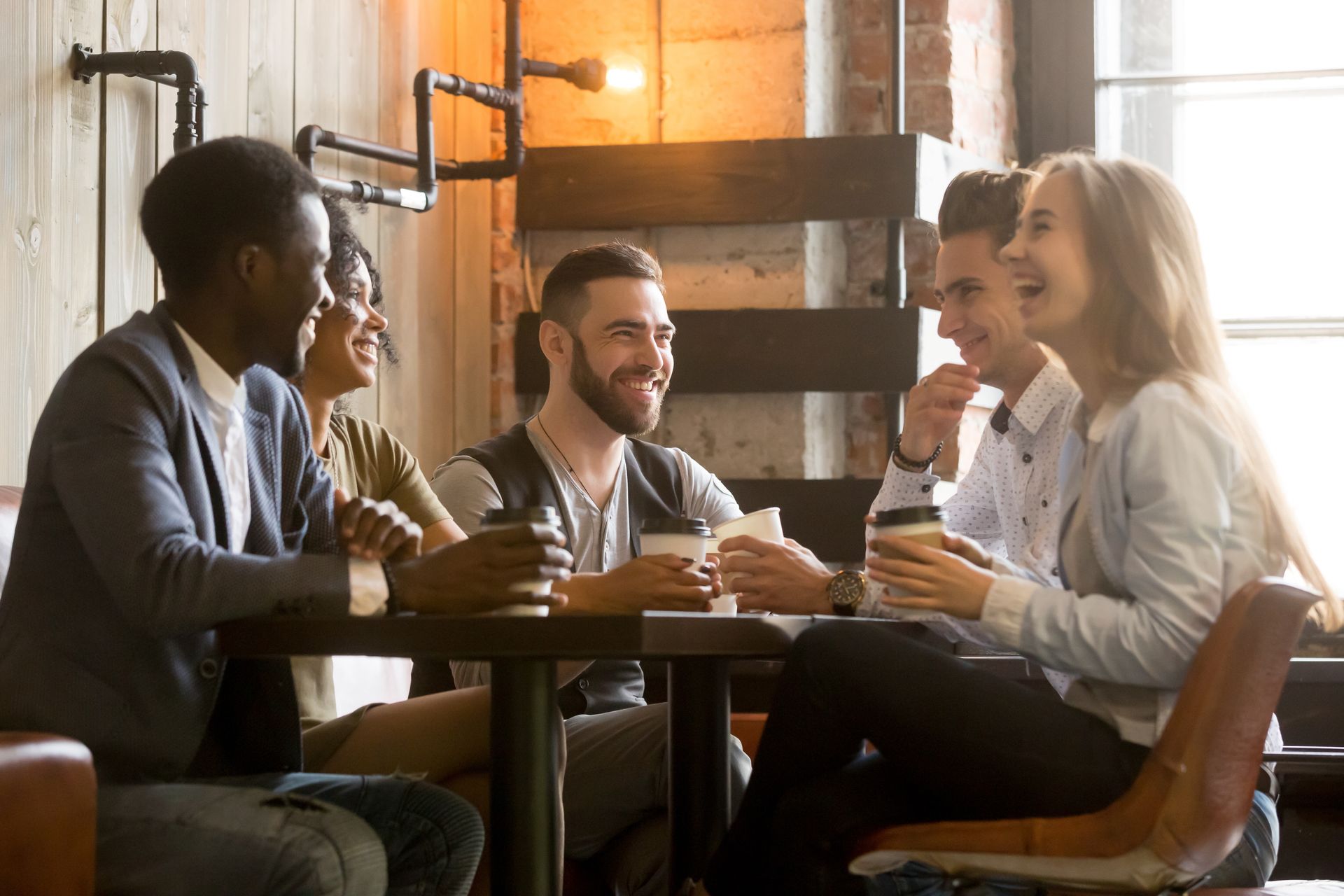 A group of people laughing and talking while holding coffee cups around a small table in a cozy, dimly lit cafe.