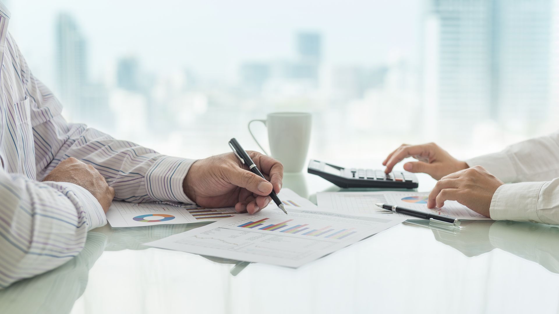 Two professionals review financial charts and data on a glass table in a bright, modern office.