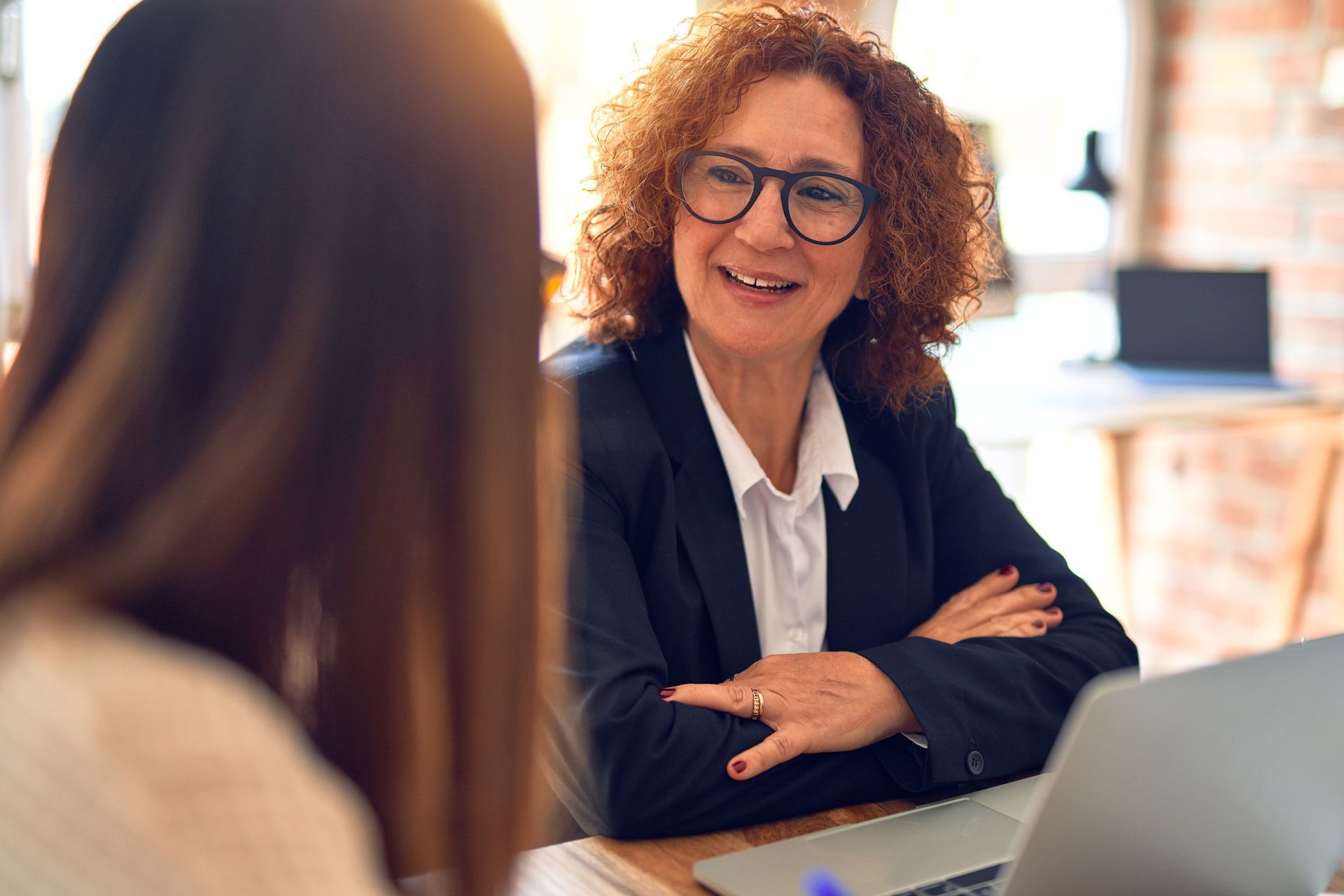 A person with curly hair and glasses smiles while talking to another person in a workspace.