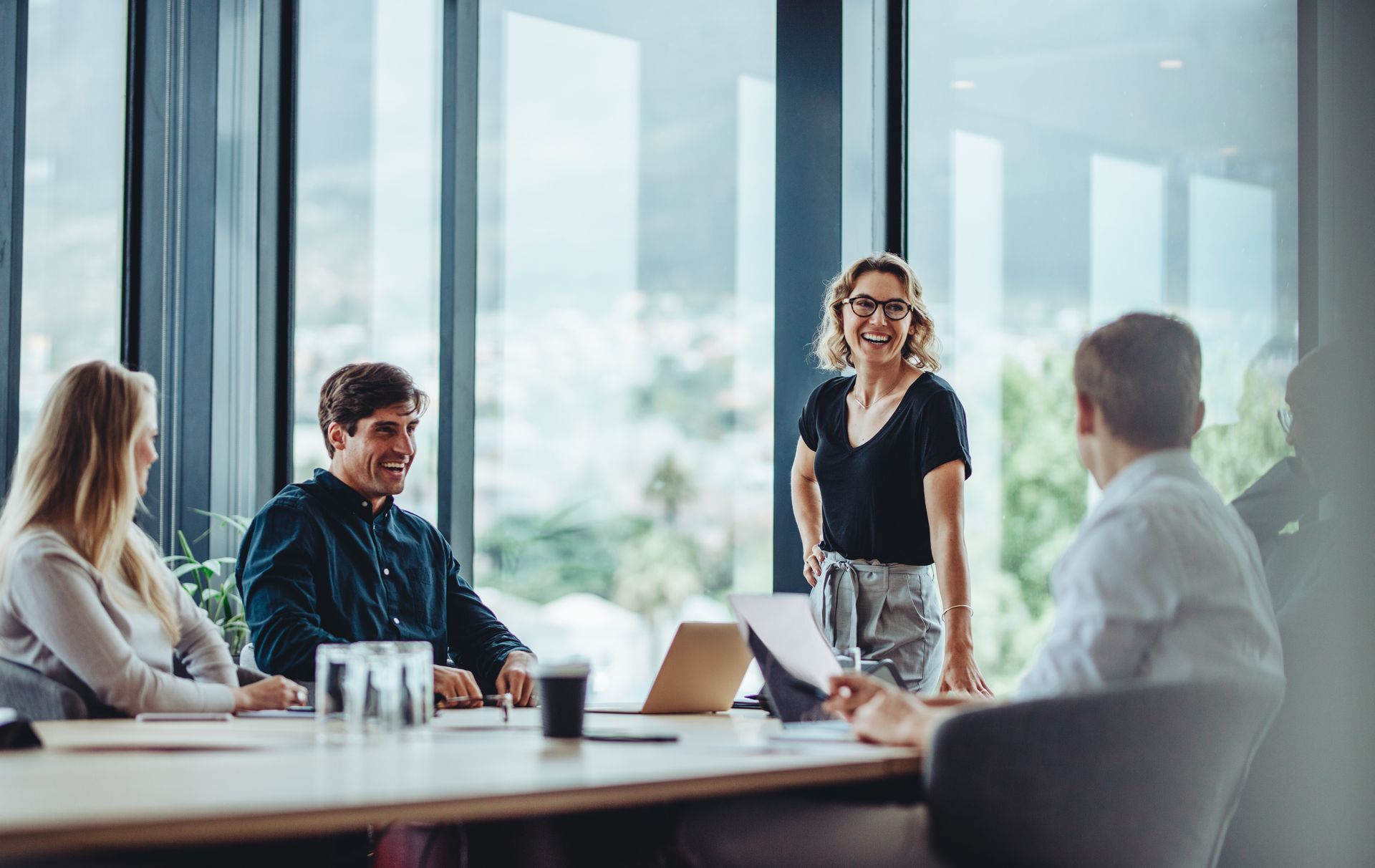 A team meeting in a sunlit office where a smiling person stands and talks to three colleagues sitting at a table.