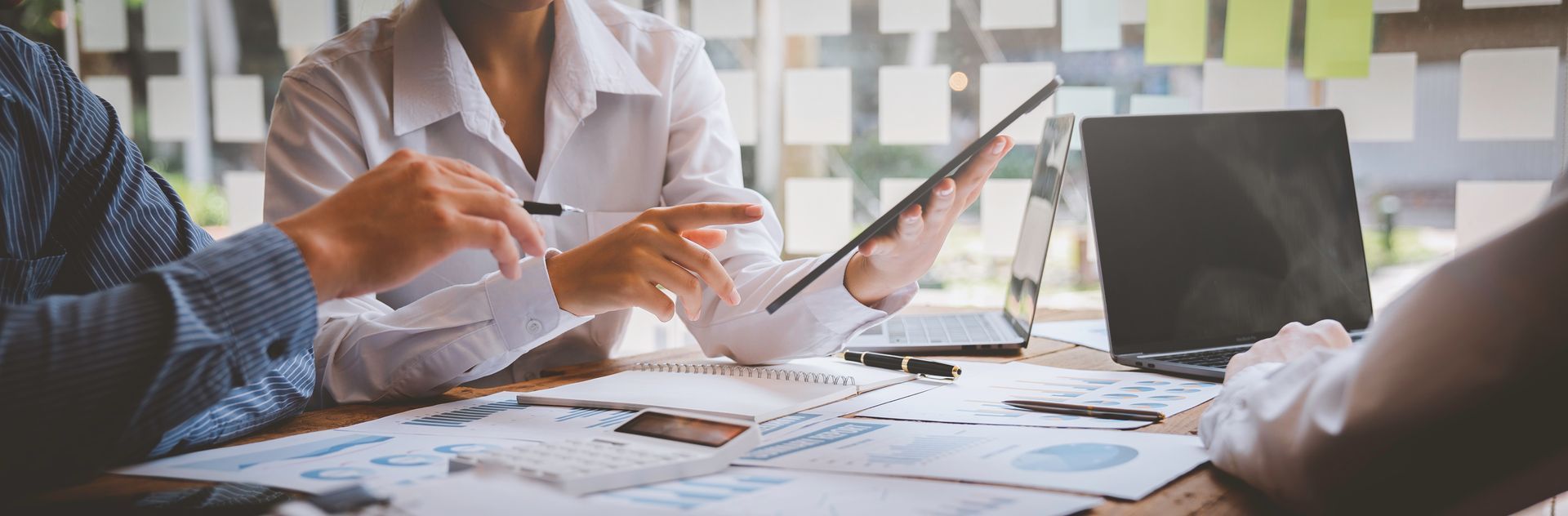Three people collaborating in a professional office, reviewing charts and data on papers and tablets at a wooden table.