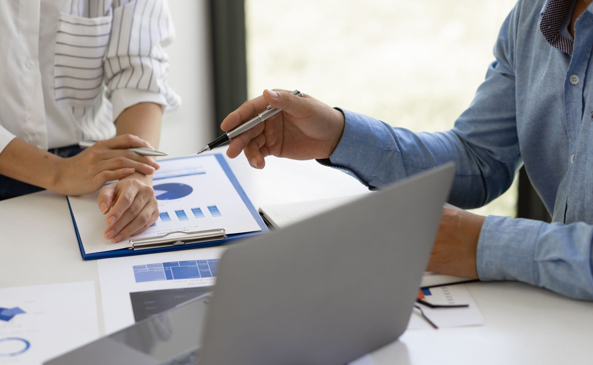 Two people discuss business charts and data on a clipboard while seated at a table with a laptop.