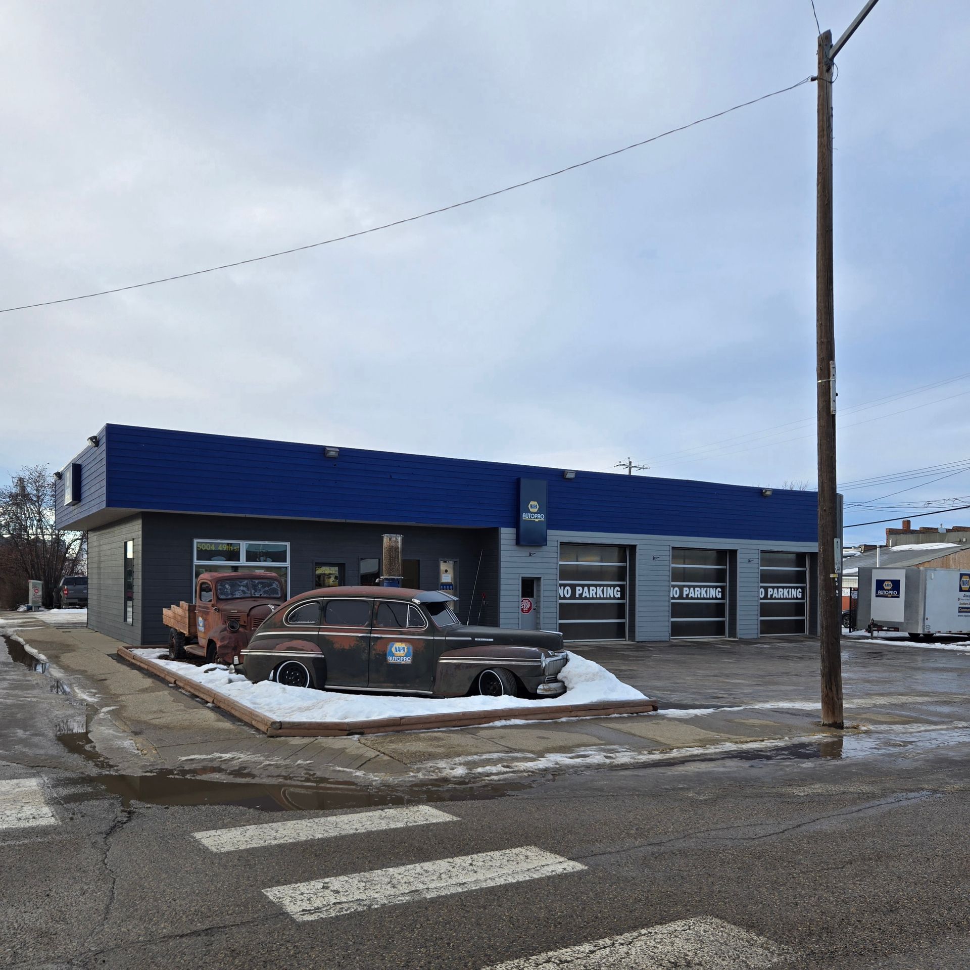 Garage exterior with blue roof and three bay doors. Two rusty cars sit in front, with a crosswalk in the foreground.