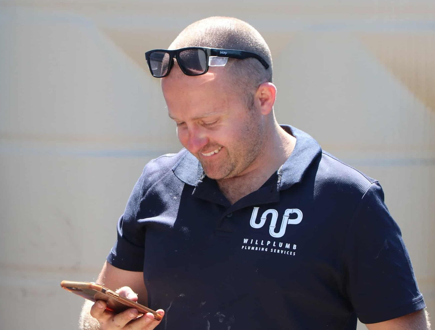 A smiling person in a dark blue polo shirt with a white logo, wearing sunglasses on their head while looking at a phone.