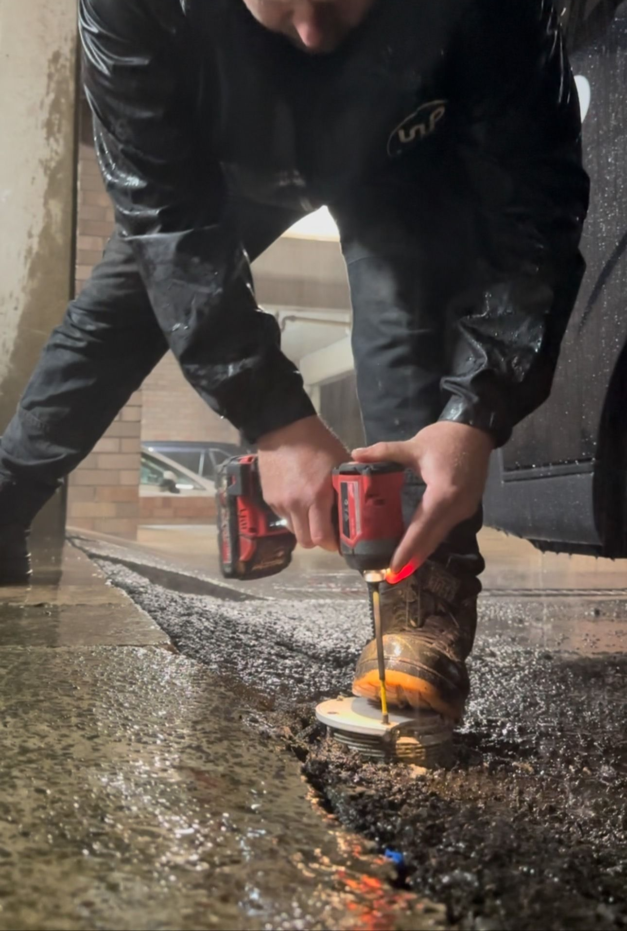 A person in a dark jacket using a red power drill to secure a circular metal bracket into wet pavement.