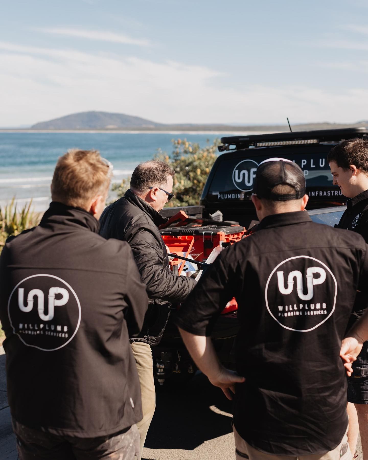 Four people in branded black uniforms stand around the back of a truck overlooking a scenic coastline.