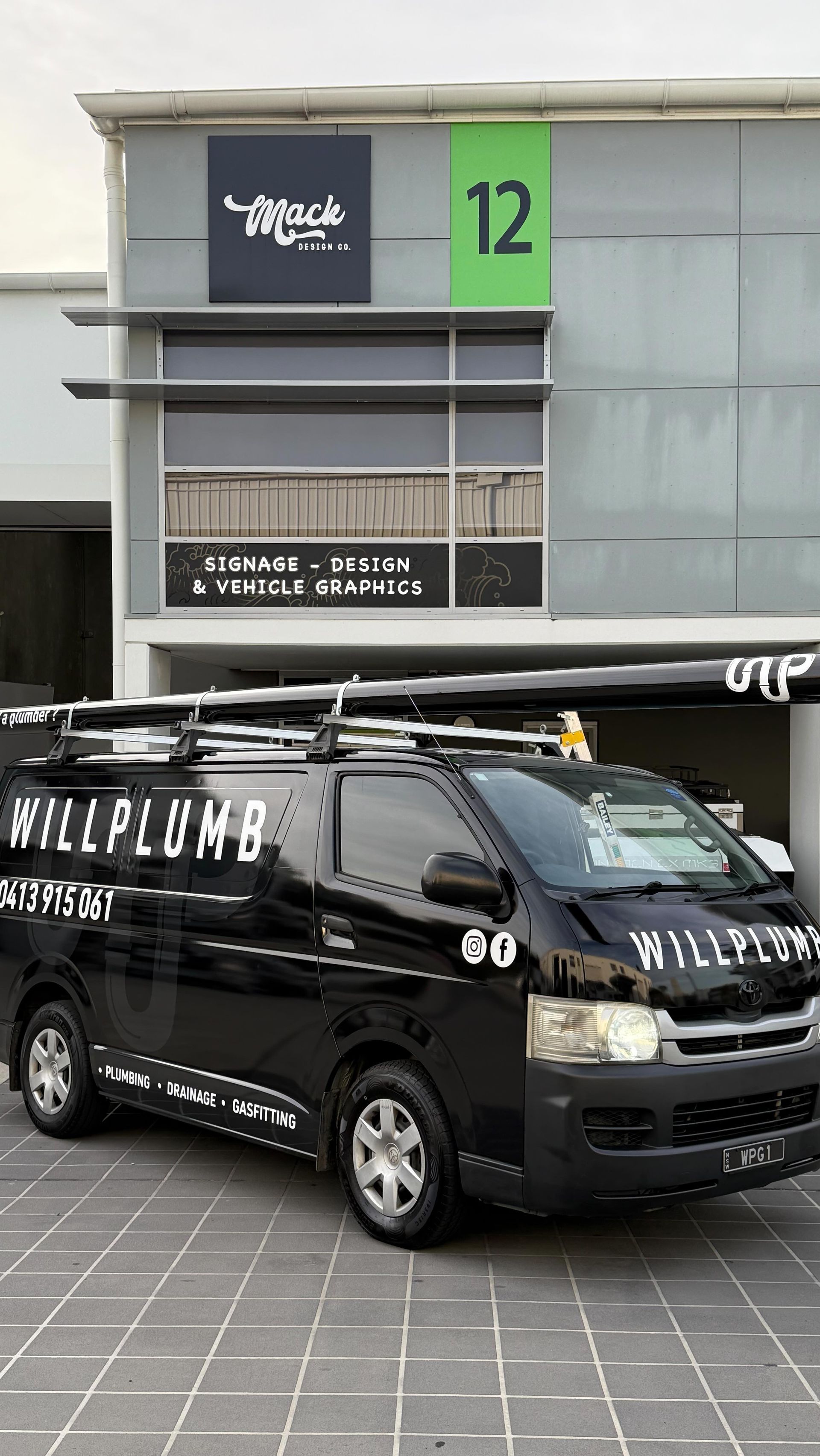 A black Willplumb work van parked in front of a commercial building labeled