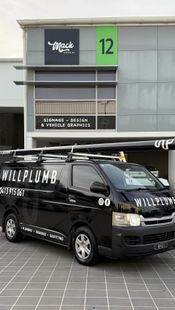 A black Willplumb work van parked in front of a commercial building labeled unit 12.