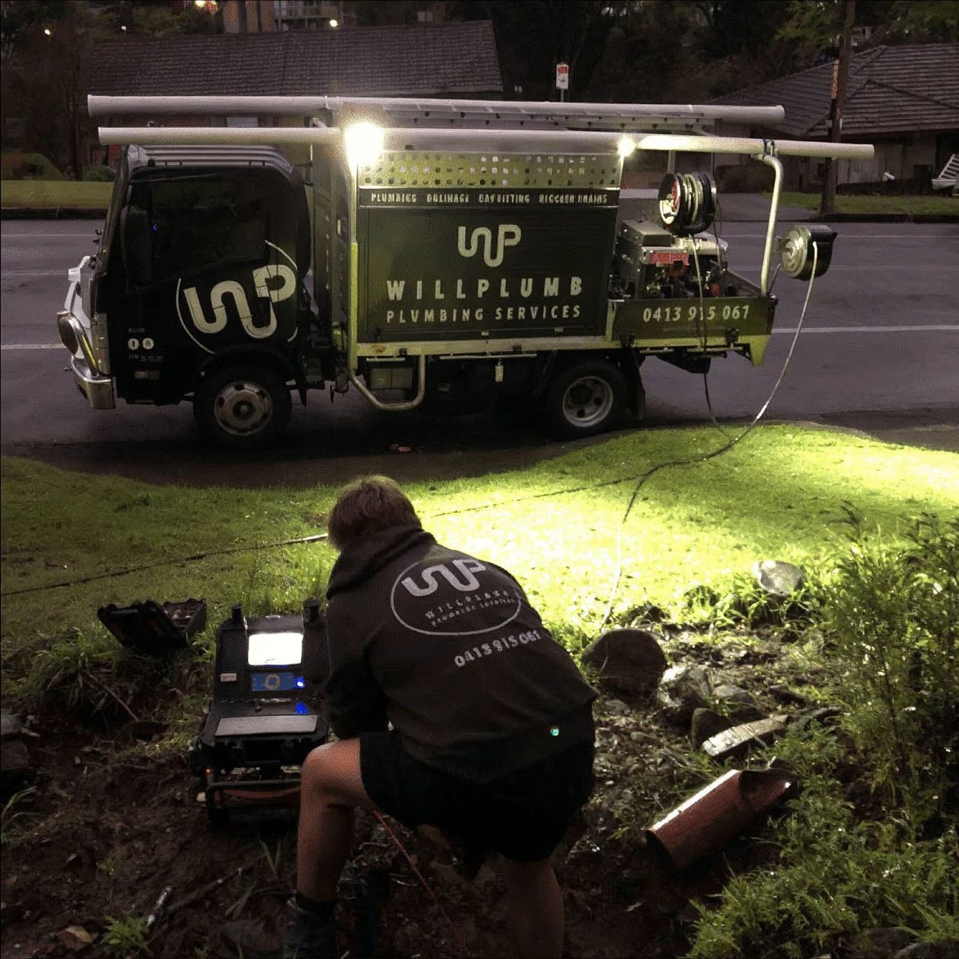 A technician uses a piece of equipment to inspect a drain at night next to a Willplumb branded work truck.