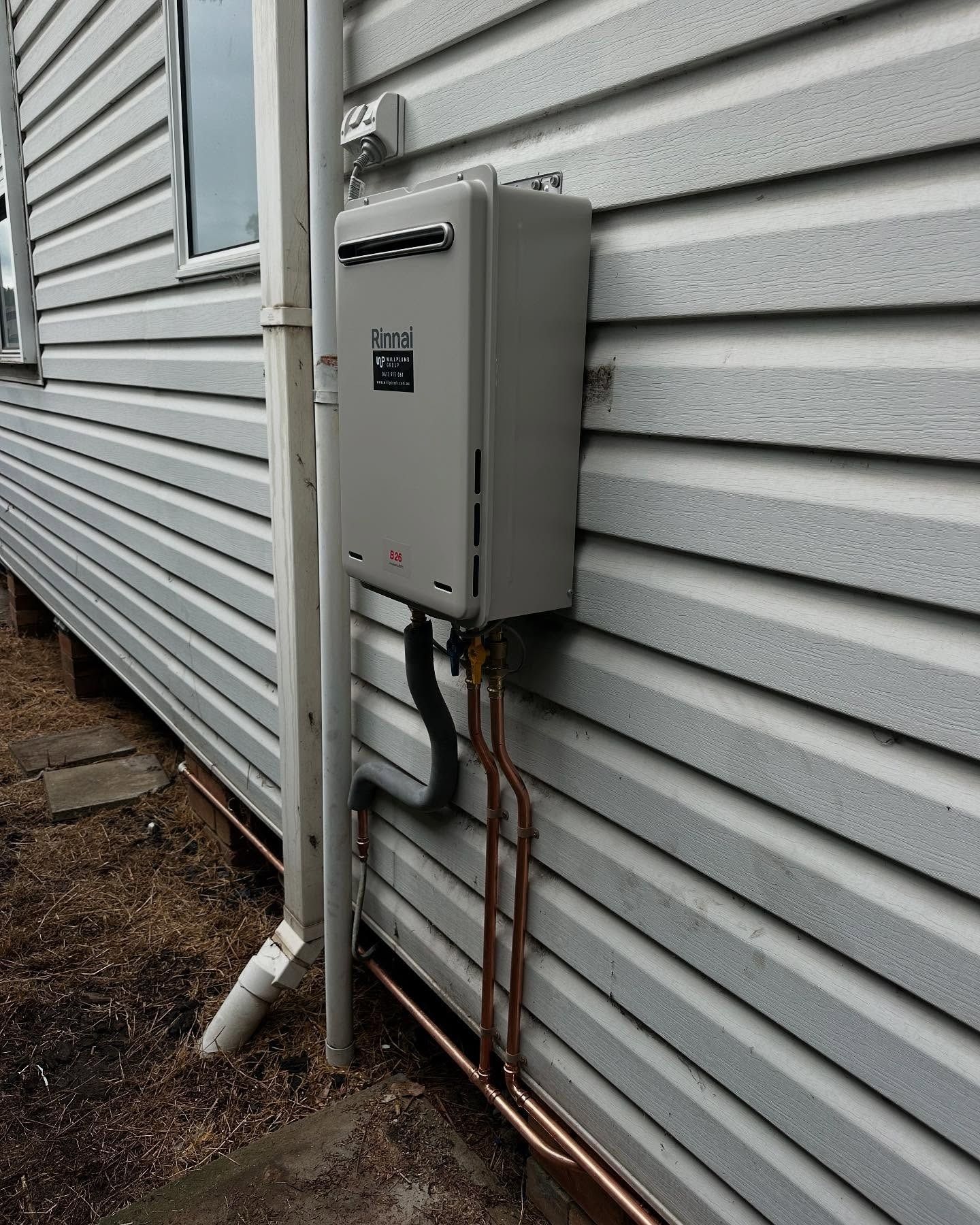A grey outdoor tankless water heater mounted on the beige vinyl siding of a house with visible copper plumbing pipes.