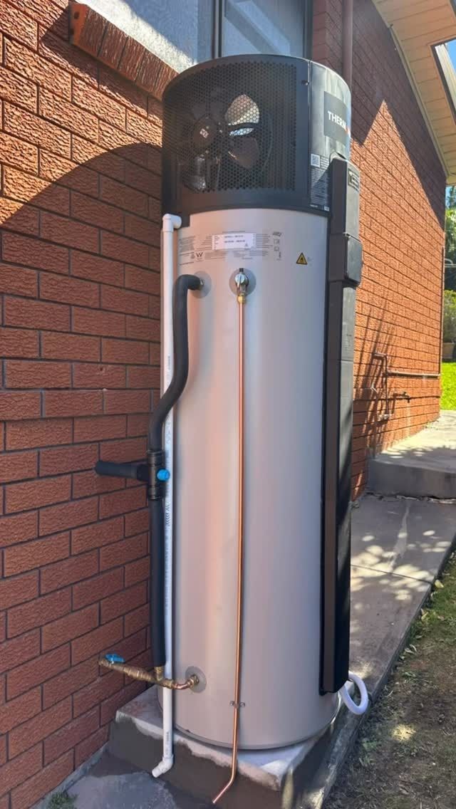 A white Thermann natural gas continuous flow water heater mounted on an outdoor red brick wall with exposed pipes.