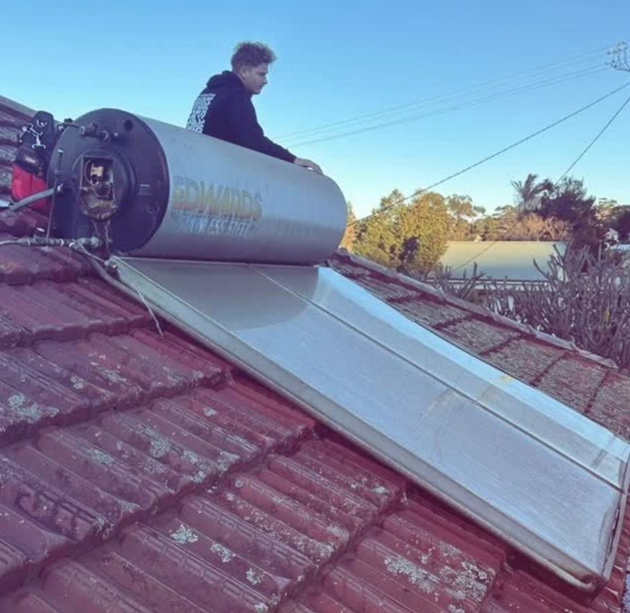 A person sits on a tiled roof beside a horizontal Edwards solar water heater unit during the day.