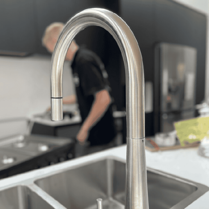 A brushed metal kitchen faucet in the foreground with a blurry person working in the background kitchen.