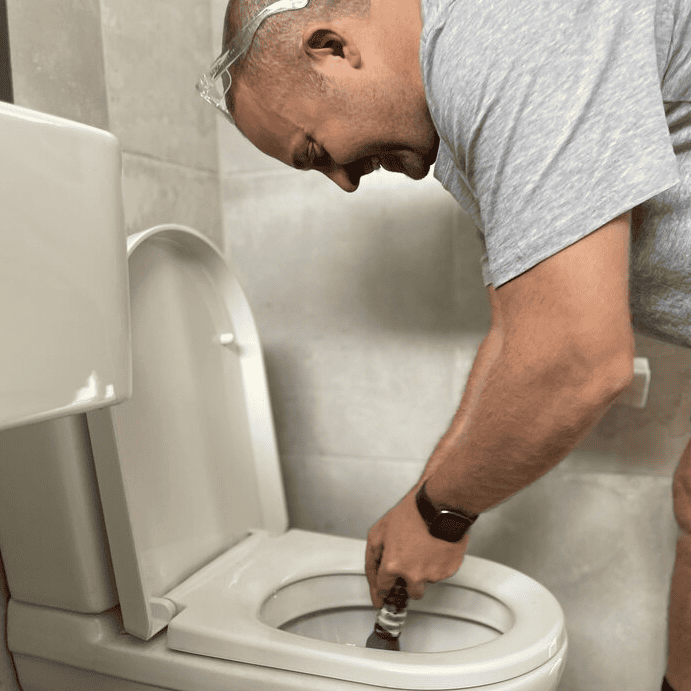 A person wearing safety glasses uses a plunger to clear a blockage in a white toilet in a tiled bathroom.